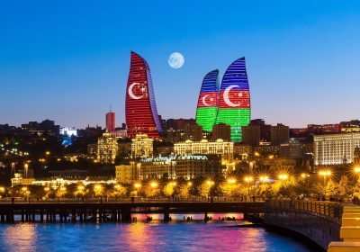 Baku skyline at night featuring the Flame Towers illuminated with the flags of Turkey and Azerbaijan, with a full moon in the background and city lights reflecting on the water.