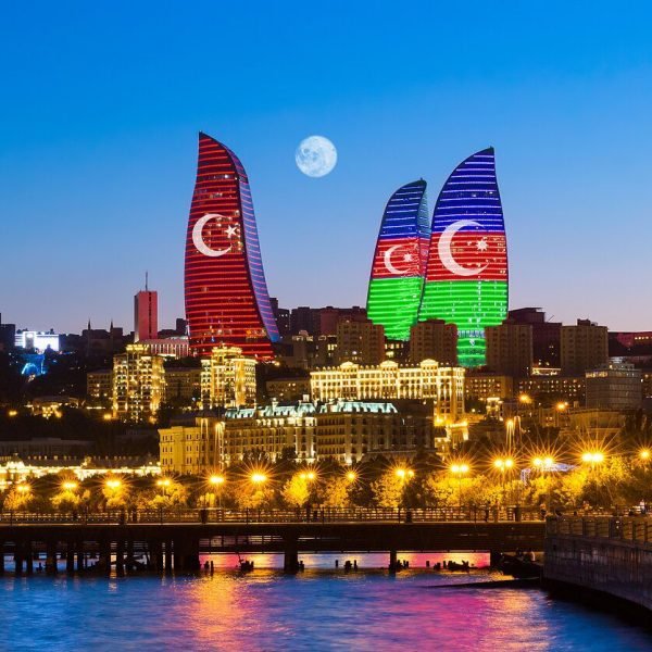 Baku skyline at night featuring the Flame Towers illuminated with the flags of Turkey and Azerbaijan, with a full moon in the background and city lights reflecting on the water.