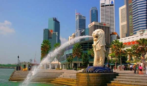 Merlion statue in Singapore with a water fountain, surrounded by modern skyscrapers and palm trees, under a clear blue sky.