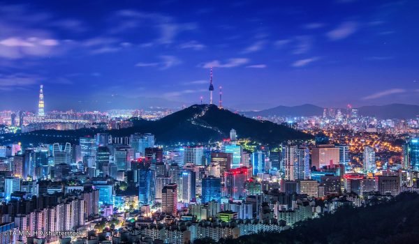 Night view of Seoul, South Korea, showcasing vibrant city lights and the iconic Namsan Tower on Namsan Mountain, with a clear blue sky in the background.