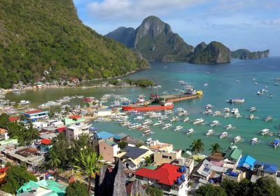Aerial view of a vibrant coastal town with colorful buildings, a busy harbor filled with boats, and lush green mountains in the background, showcasing the scenic beauty of the area.
