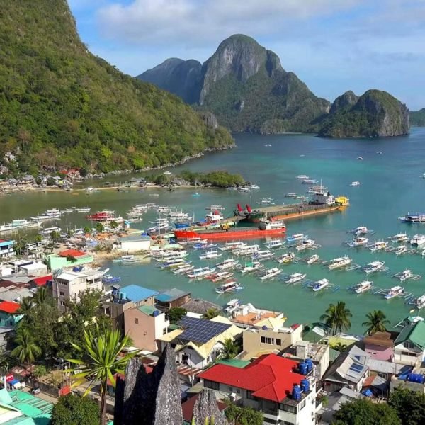 Aerial view of a vibrant coastal town with colorful buildings, a busy harbor filled with boats, and lush green mountains in the background, showcasing the scenic beauty of the area.