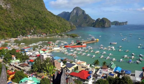 Aerial view of El Nido, Philippines, showcasing a vibrant coastal town with colorful buildings, a busy harbor filled with boats, and stunning limestone cliffs in the background against a clear blue sky.