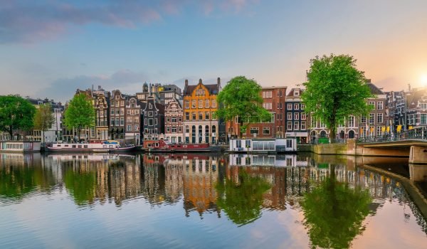 Scenic view of Amsterdam's historic canals lined with colorful buildings and lush green trees, reflecting in the water during sunset.