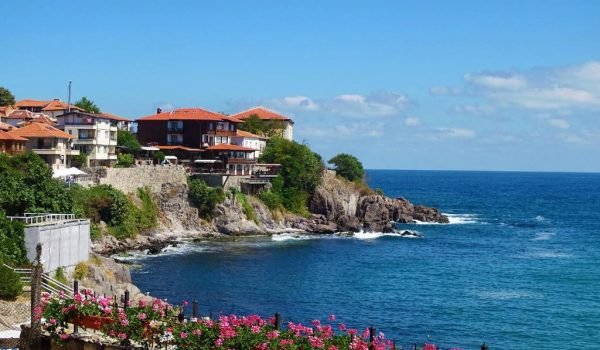 Coastal view of a seaside town with colorful houses and blooming flowers along the shore, overlooking a vibrant blue ocean under a clear sky.