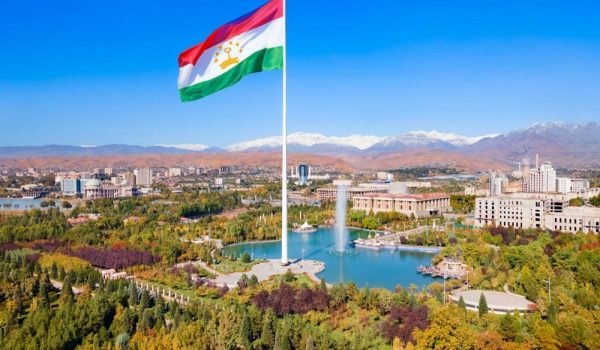 Aerial view of a large Tajikistan flag flying over a scenic park with a lake and mountains in the background, showcasing the natural beauty and urban landscape of Dushanbe, Tajikistan.