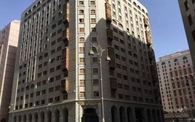 Modern hotel building in a bustling urban area, featuring a blend of architectural styles with prominent windows and decorative elements, surrounded by other structures and street activity. Ideal for travelers seeking accommodation in a central location.