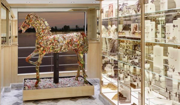 Artistic horse sculpture made from colorful materials displayed in a modern gallery, alongside a glass case showcasing various intricate art pieces and collectibles.