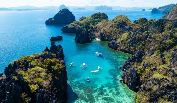 Aerial view of a tropical lagoon surrounded by lush green islands and clear turquoise waters, with boats anchored in the bay, showcasing the natural beauty of a remote coastal paradise.