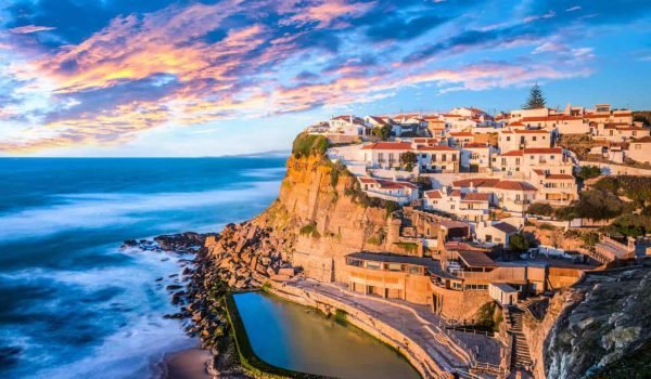 Scenic coastal view of a cliffside village at sunset, showcasing colorful buildings, rocky shoreline, and a tranquil pool reflecting the vibrant sky.