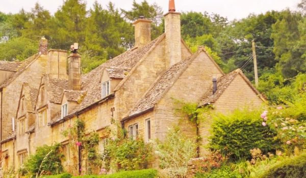 Charming stone cottage surrounded by lush greenery, featuring a sloped roof, multiple chimneys, and colorful garden plants in the Cotswolds.