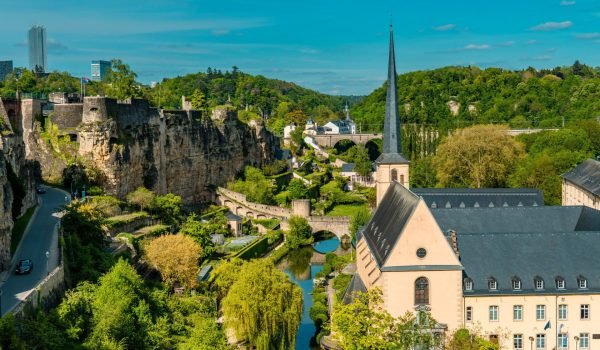 Scenic view of Luxembourg City showcasing the historic church with a tall spire, surrounded by lush greenery and a winding river, highlighting the picturesque landscape and architectural beauty of the region.