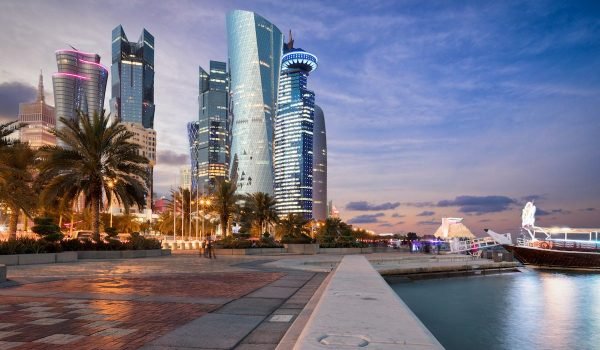 Modern skyline of a city with illuminated skyscrapers at dusk, featuring palm trees along a waterfront promenade.