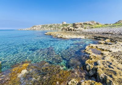 Scenic coastal view featuring clear turquoise waters and rocky shoreline, with lush greenery and historical structures in the background under a bright blue sky.