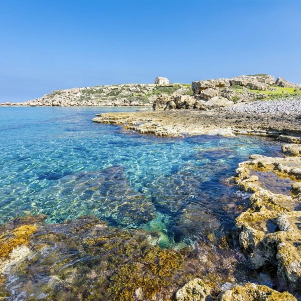 Scenic coastal view featuring clear turquoise waters and rocky shoreline, with lush greenery and historical structures in the background under a bright blue sky.