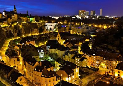 Aerial view of Luxembourg City at night, showcasing illuminated historic buildings and modern skyscrapers against a twilight sky.