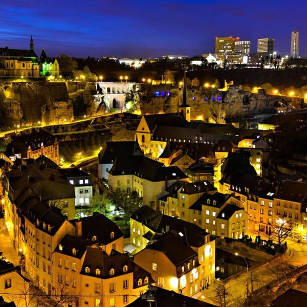 Aerial view of Luxembourg City at night, showcasing illuminated historic buildings and modern skyscrapers against a twilight sky.