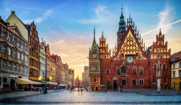 Historic architecture of the Main Market Square in Gdańsk, Poland, featuring the iconic Gothic-style Town Hall and colorful merchant houses under a vibrant sunset sky.