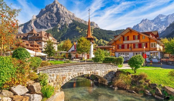 Scenic view of a charming village with traditional architecture, a stone bridge over a clear river, surrounded by lush greenery and majestic mountains under a bright blue sky.