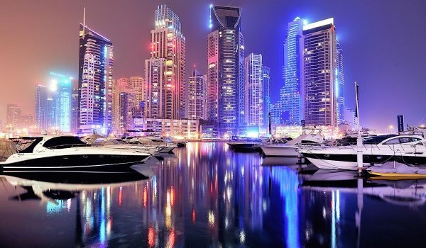 Nighttime cityscape of modern skyscrapers illuminated with colorful lights, reflecting on a calm harbor filled with boats.