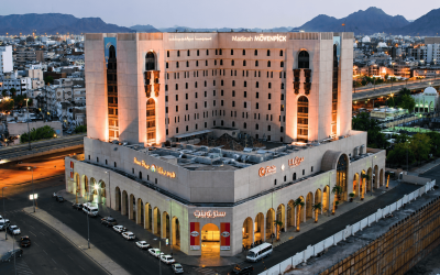 Madinah Mövenpick Hotel exterior view at dusk, showcasing its modern architecture and illuminated signage, surrounded by urban landscape and mountains in the background.
