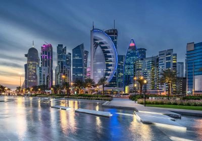 Cityscape of Doha, Qatar, showcasing modern skyscrapers illuminated at dusk, with the iconic Aspire Tower in the foreground and reflections on the water.