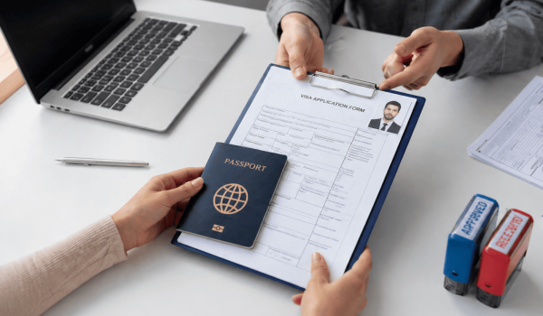 Person submitting a visa application form alongside a passport at a desk with a laptop and staplers in the background.