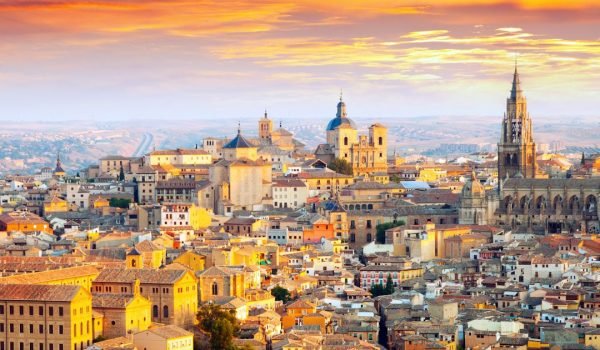 Aerial view of Toledo, Spain, showcasing the historic architecture and vibrant rooftops under a colorful sunset sky.