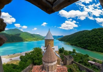 Scenic view of a medieval church overlooking a turquoise lake and lush green mountains under a blue sky with fluffy clouds.