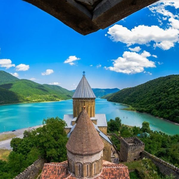 Scenic view of a medieval church overlooking a turquoise lake and lush green mountains under a blue sky with fluffy clouds.