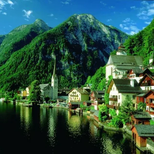 Scenic view of a picturesque lakeside village surrounded by lush green mountains, featuring charming houses and a church steeple, reflecting in the calm water under a clear blue sky.