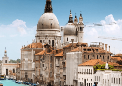 Scenic view of the Grand Canal in Venice, featuring historic buildings with domed roofs and terracotta tiles, under a clear blue sky, showcasing the city's iconic architecture and waterways.