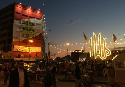Crowd enjoying an outdoor festival at sunset, with colorful lights adorning buildings and decorations in the background.