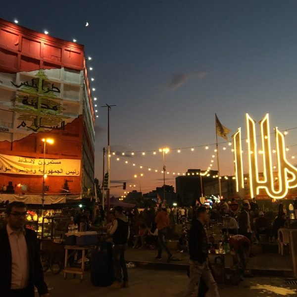 Crowd enjoying an outdoor festival at sunset, with colorful lights adorning buildings and decorations in the background.