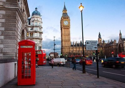 Red telephone booth near Big Ben in London, with busy street traffic and historic buildings in the background during twilight.