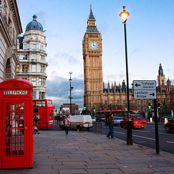 Red telephone booth near Big Ben in London, with busy street traffic and historic buildings in the background during twilight.