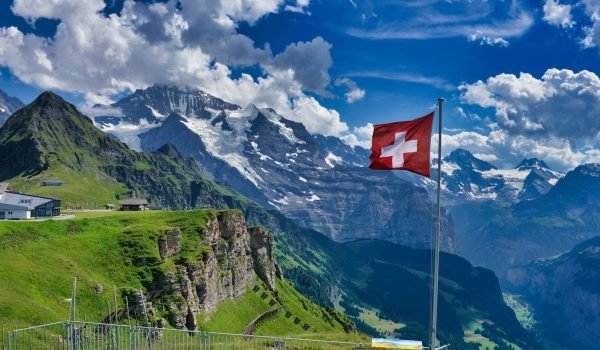 Swiss flag waving in front of majestic mountains under a cloudy blue sky, showcasing the natural beauty of Switzerland.