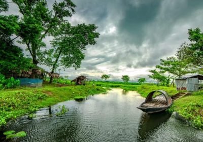 Scenic landscape featuring a tranquil river surrounded by lush greenery and trees, with traditional huts in the background under a dramatic cloudy sky.