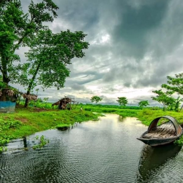 Scenic landscape featuring a tranquil river surrounded by lush greenery and trees, with traditional huts in the background under a dramatic cloudy sky.