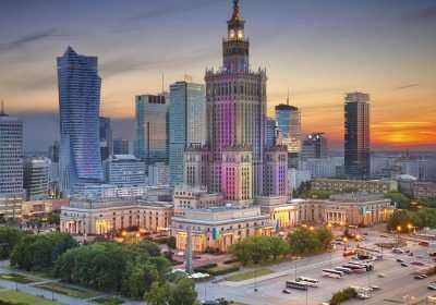 Panoramic view of Warsaw, Poland, featuring the iconic Palace of Culture and Science illuminated at sunset, surrounded by modern skyscrapers and bustling city life.