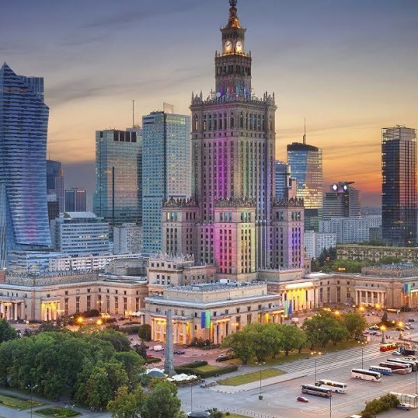 Panoramic view of Warsaw, Poland, featuring the iconic Palace of Culture and Science illuminated at sunset, surrounded by modern skyscrapers and bustling city life.