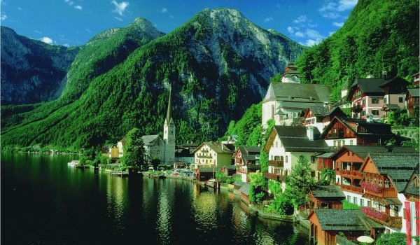 Scenic view of a picturesque village by a lake, surrounded by lush green mountains and clear blue skies, showcasing traditional architecture and a serene landscape.