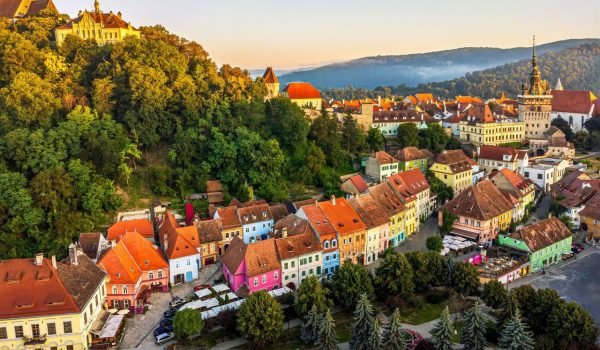 Aerial view of a picturesque town with colorful houses and a castle on a hill, surrounded by lush greenery and misty mountains in the background during sunrise.