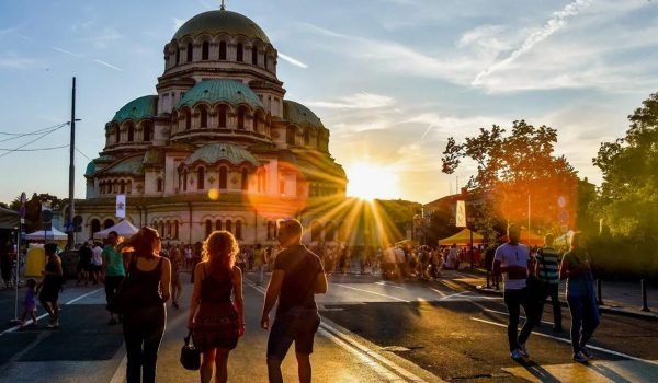 Sunset view of the Alexander Nevsky Cathedral in Sofia, Bulgaria, with silhouettes of people walking on a street, creating a vibrant atmosphere.