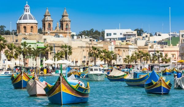 Colorful traditional fishing boats in a vibrant harbor with palm trees and historic buildings in the background, showcasing the scenic beauty of Malta's waterfront.