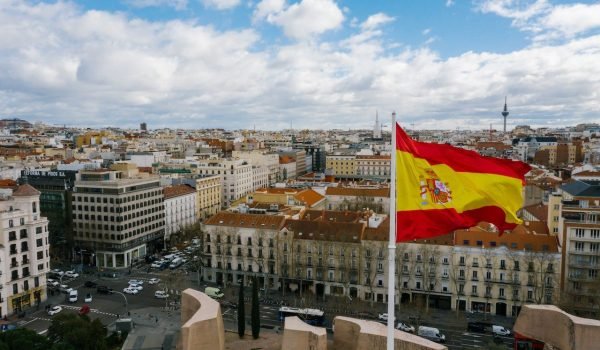 Aerial view of Madrid, Spain, showcasing the city skyline with the Spanish flag prominently waving in the foreground against a backdrop of blue skies and scattered clouds.