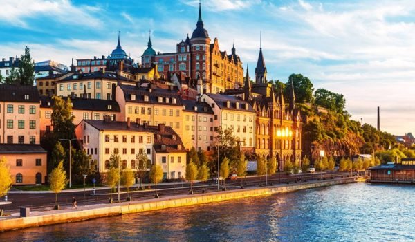 Scenic view of a historic castle overlooking a river at sunset, featuring colorful buildings and lush greenery along the waterfront.