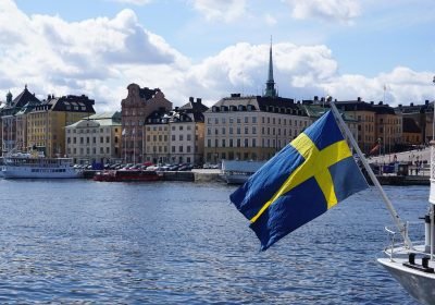 Swedish flag waving in the foreground with a picturesque view of a waterfront city, featuring historic buildings and a church spire under a partly cloudy sky.