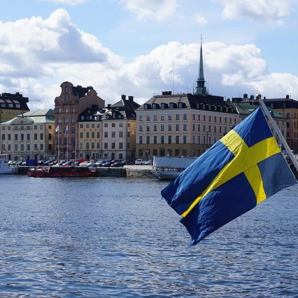 Swedish flag waving in the foreground with a picturesque view of a waterfront city, featuring historic buildings and a church spire under a partly cloudy sky.