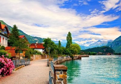 Scenic lakeside pathway with lush greenery, mountains in the background, and a clear blue sky, perfect for leisurely walks and nature photography.